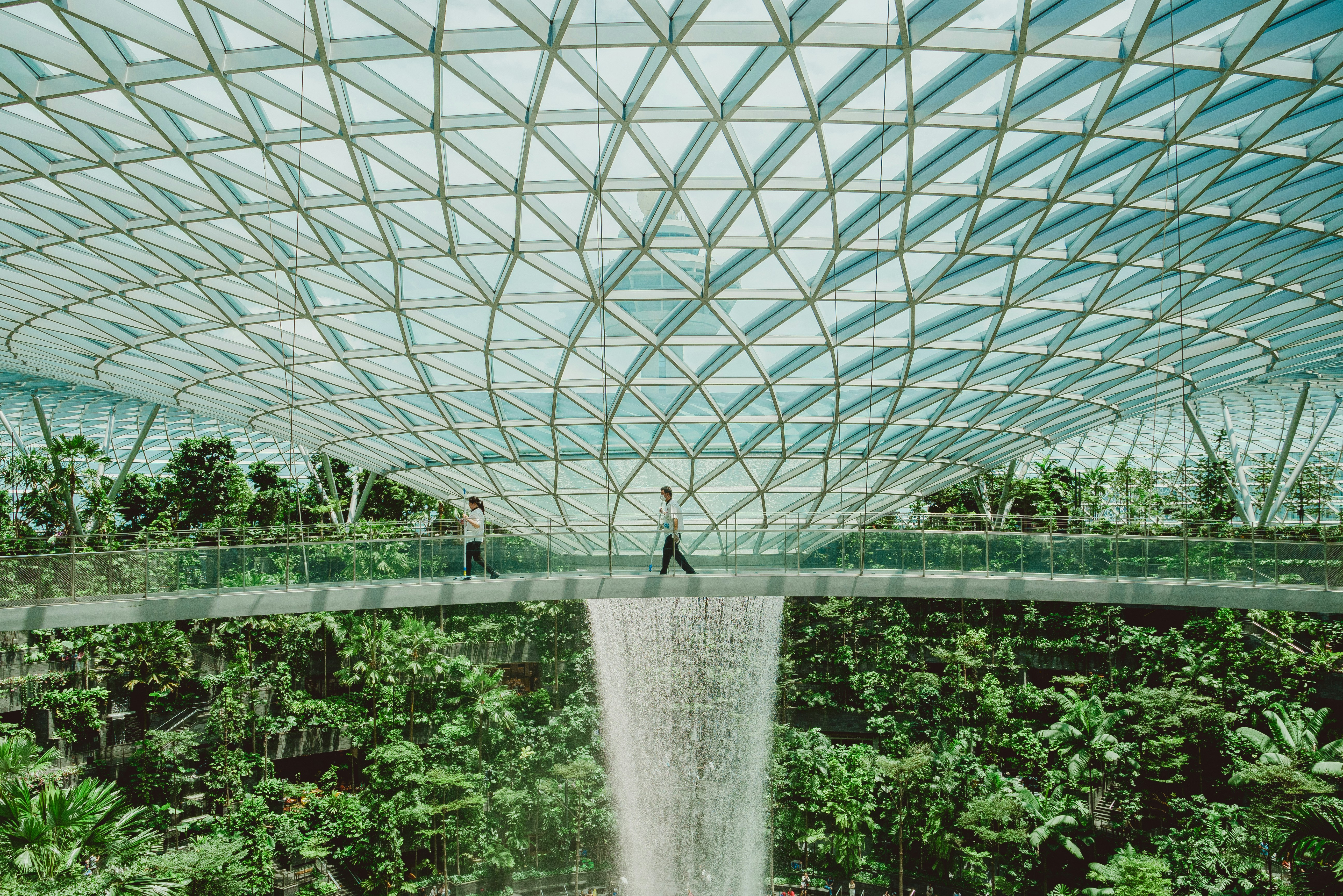 hanna-lazar-biodome-unsplash.jpg Top of a biodome, showing a glass ceiling releasing a waterfall, with a walkway used by two people walking, and a teeming mass of green growing plants below