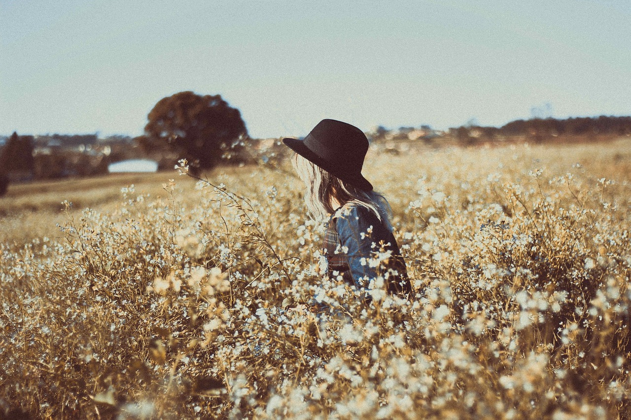 A girl in a sunny field, with a slough in the background, wearing a fedora