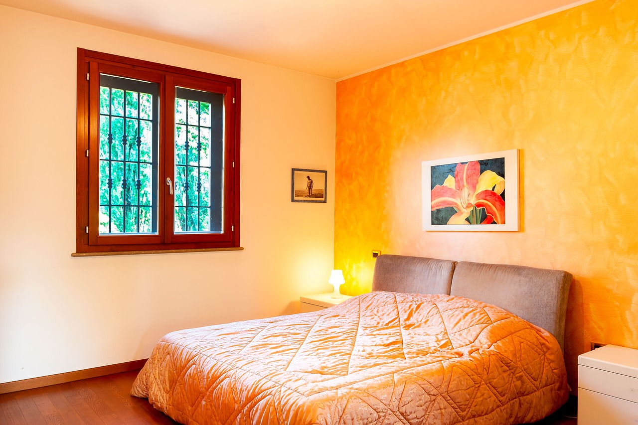 A bedroom in an orange theme, with orange walls, an bed with an orange bedspread, an orange flower painting on the wall, and a window framed in orangish arbutus wood