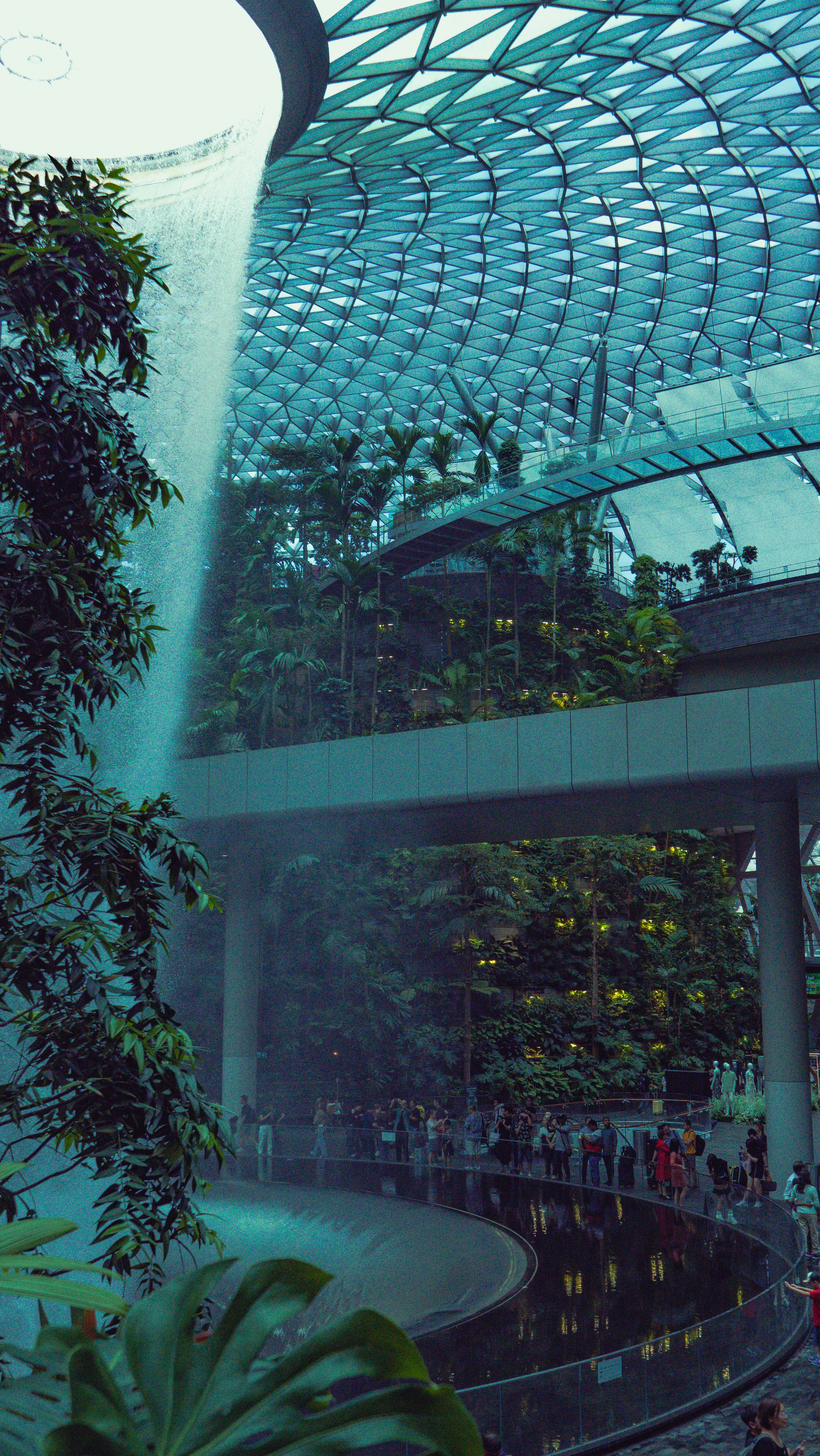 hanna-lazar-kVx5YHZLtfU-unsplash.jpg An interior view of a biodome of teeming green and fruiting plants, with a waterfall cascading into a pool, and the walkway near the pool is lined with people